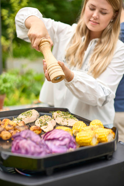 A person uses a wooden pepper mill to season chicken, corn, potatoes, and red cabbage cooking on an outdoor grill pan.