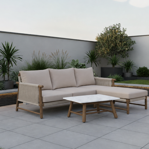 A modern outdoor sectional sofa with beige cushions and a matching ottoman, paired with a small rectangular coffee table, set on a light gray tiled patio in a landscaped backyard with plants and a low wall.