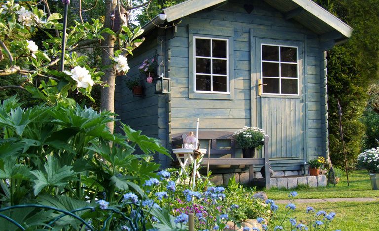 Pale blue woodpaneled summer house in a garden with plants and flowers in forefront