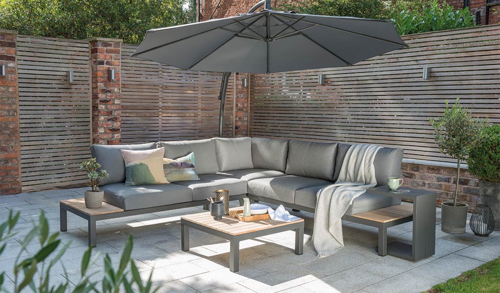 The Elba Corner Set and Elba Side Table and free arm parasol on a patio in a garden.