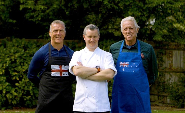 Cricketers Jeff Thomson and Alec Stewart wearing BBQ aprons, with Fat Duck chef, Ashley Hatton stood between them outdoors.