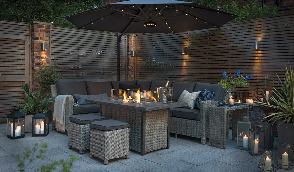 The Palma Corner Sofa with a lit Palma Fire Pit Table and 3.5m Free Arm Parasol with LED Lighting and Wireless Speaker on a patio at night.