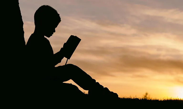 silhouette of child reading a book whilst sat leaning against a tree